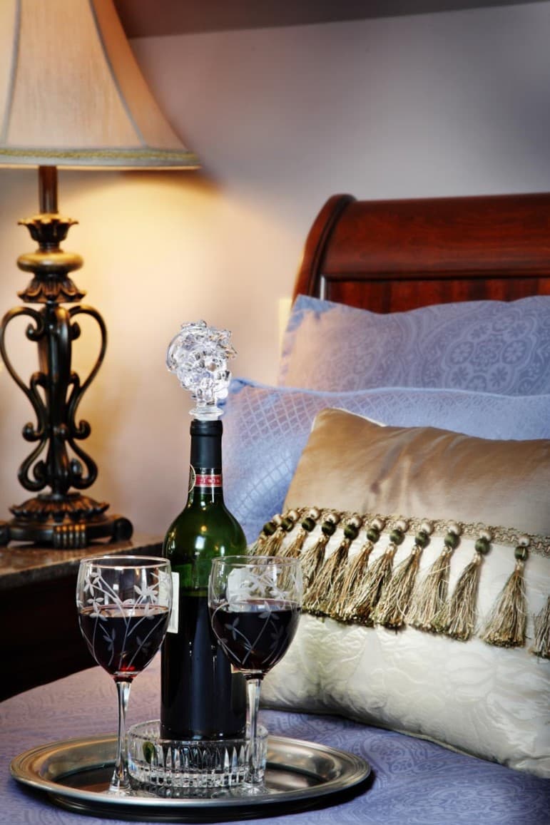 A bottle of red wine with two glasses sits on a silver tray beside a lamp and plush bedding.