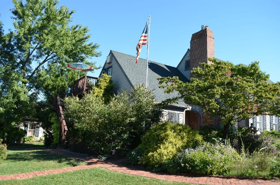 A charming house with a green roof, featuring American flag and lush surrounding gardens.