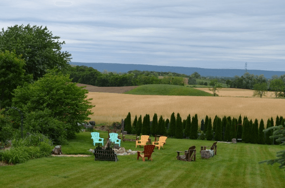 A tranquil outdoor space with colorful chairs arranged on a green lawn, set against a backdrop of rolling fields and cloudy skies.